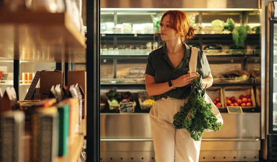 a young woman is browsing grocery store looking for fresh produce, she is carrying a bag with greens spilling out