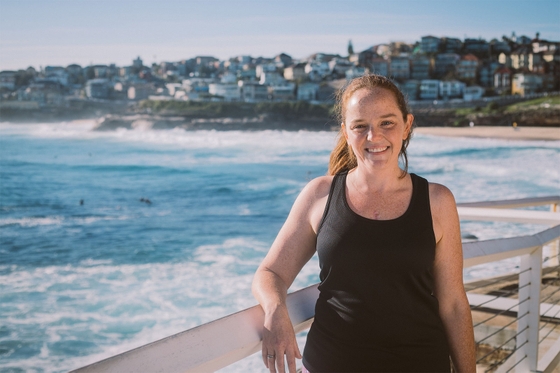 A woman in a black tank top stands on a boardwalk, gazing at the vast ocean ahead.