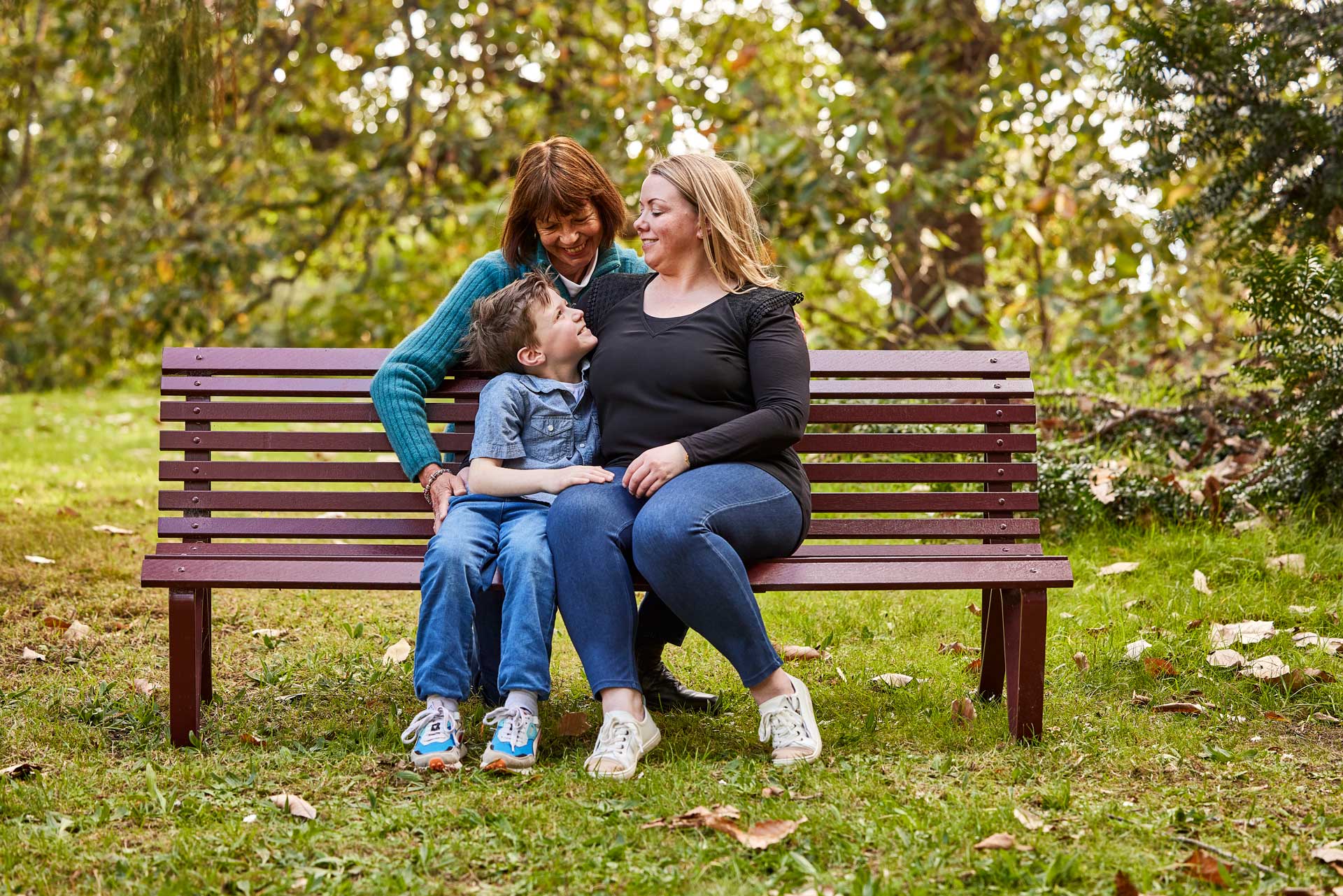 Family of three, older mum, daughter and son/grandson, sitting outdoors in a leafy park on park bench