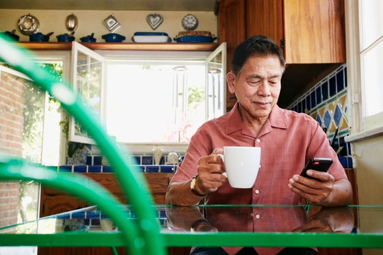 An older man sitting at his kitchen table, looking down at his smartphone in one hand and holding a mug in the other.