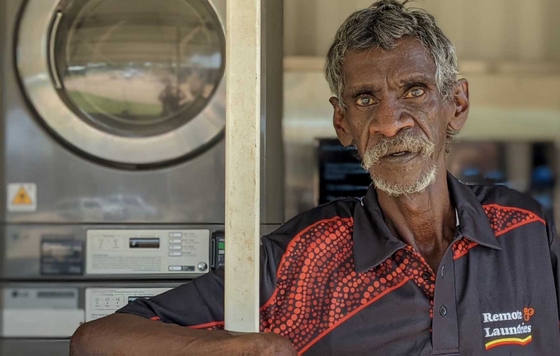 A First Nations man wearing a red shirt is positioned in front of a washing machine.