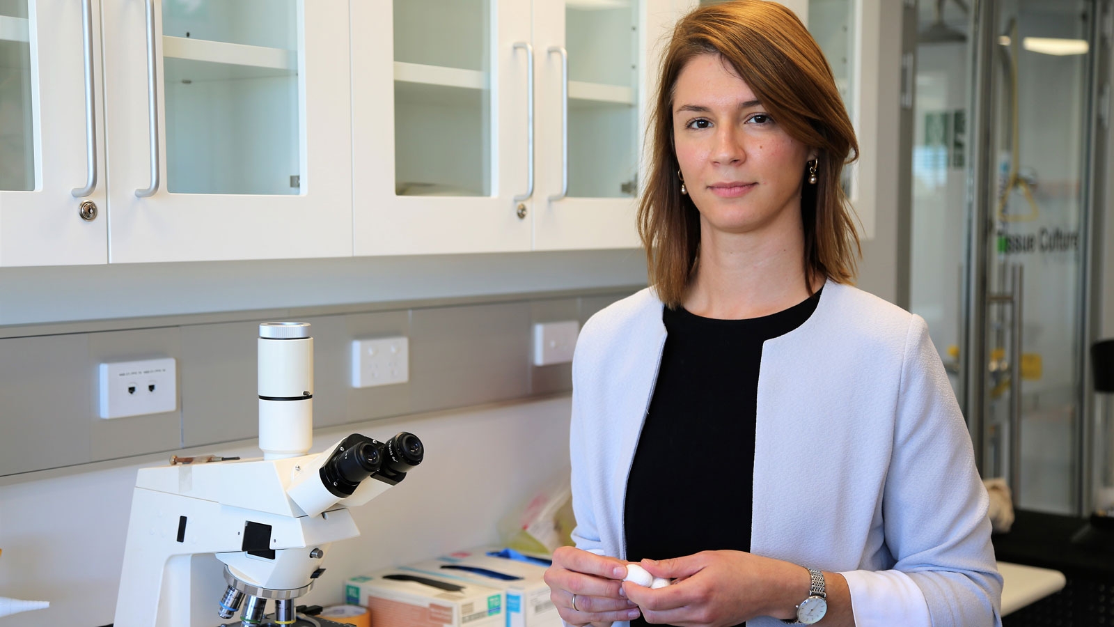 A female scientist in a lab coat stands beside a microscope, conducting research in a laboratory setting.